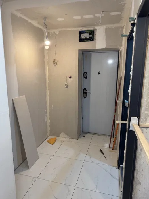 Wnętrze w trakcie prac wykończeniowych: białe płytki podłogowe, częściowo wyszpachlowane ściany, widoczna instalacja elektryczna i ościeżnica drzwi.