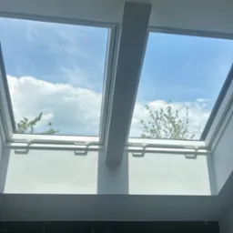 Dwa okna dachowe z widokiem na błękitne niebo i chmury, białe ramy okienne, marmurowe wykończenie ściany na dole kadru, jasne, naturalne oświetlenie.