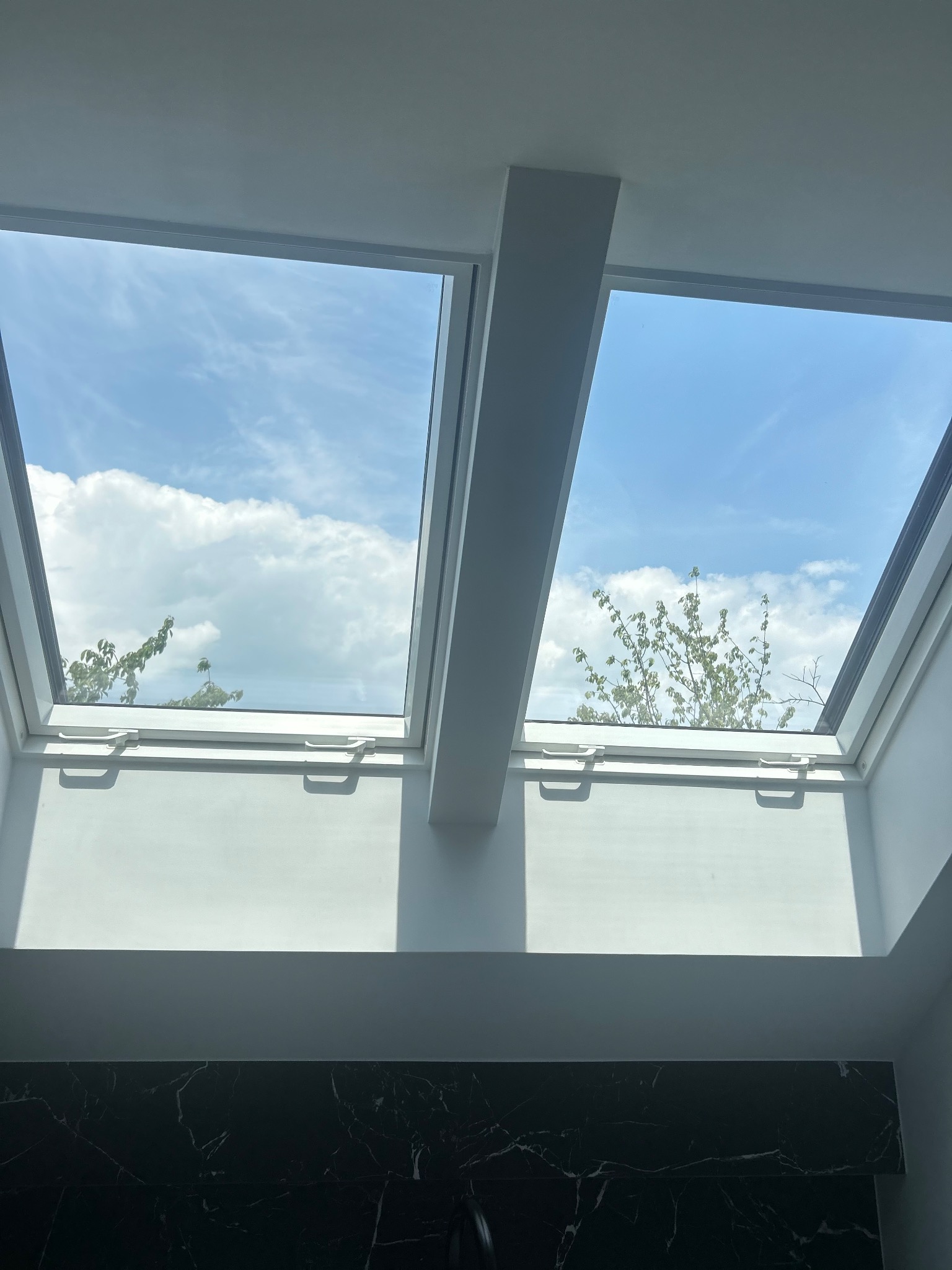 Dwa okna dachowe z widokiem na błękitne niebo i chmury, białe ramy okienne, marmurowe wykończenie ściany na dole kadru, jasne, naturalne oświetlenie.