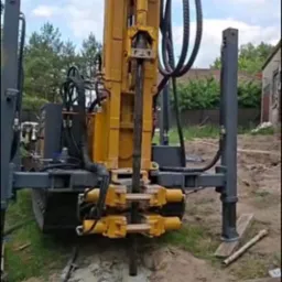 Żółta wiertnica w trakcie pracy, wiertło zagłębione w ziemi. Widoczny fragment wykopu wypełnionego płynem. W tle zabudowania i zieleń.