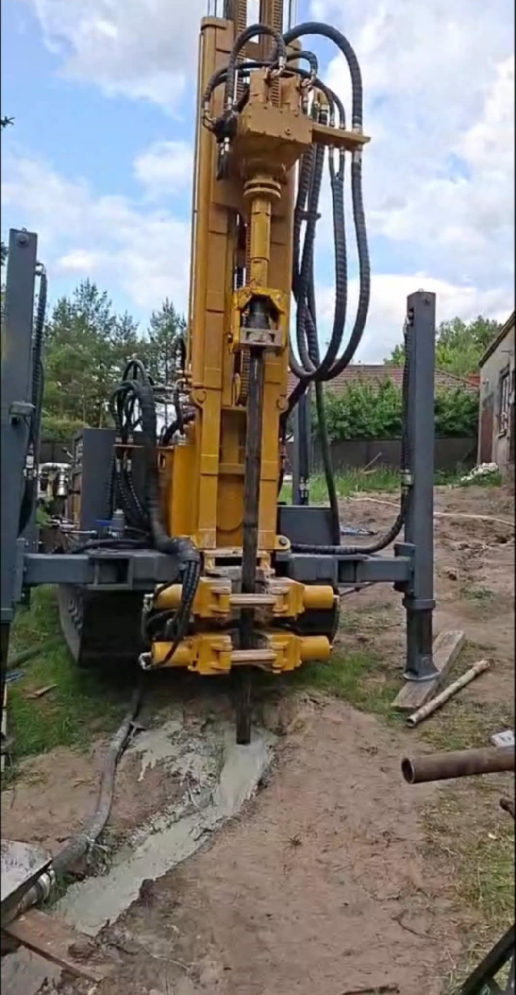 Żółta wiertnica w trakcie pracy, wiertło zagłębione w ziemi. Widoczny fragment wykopu wypełnionego płynem. W tle zabudowania i zieleń.