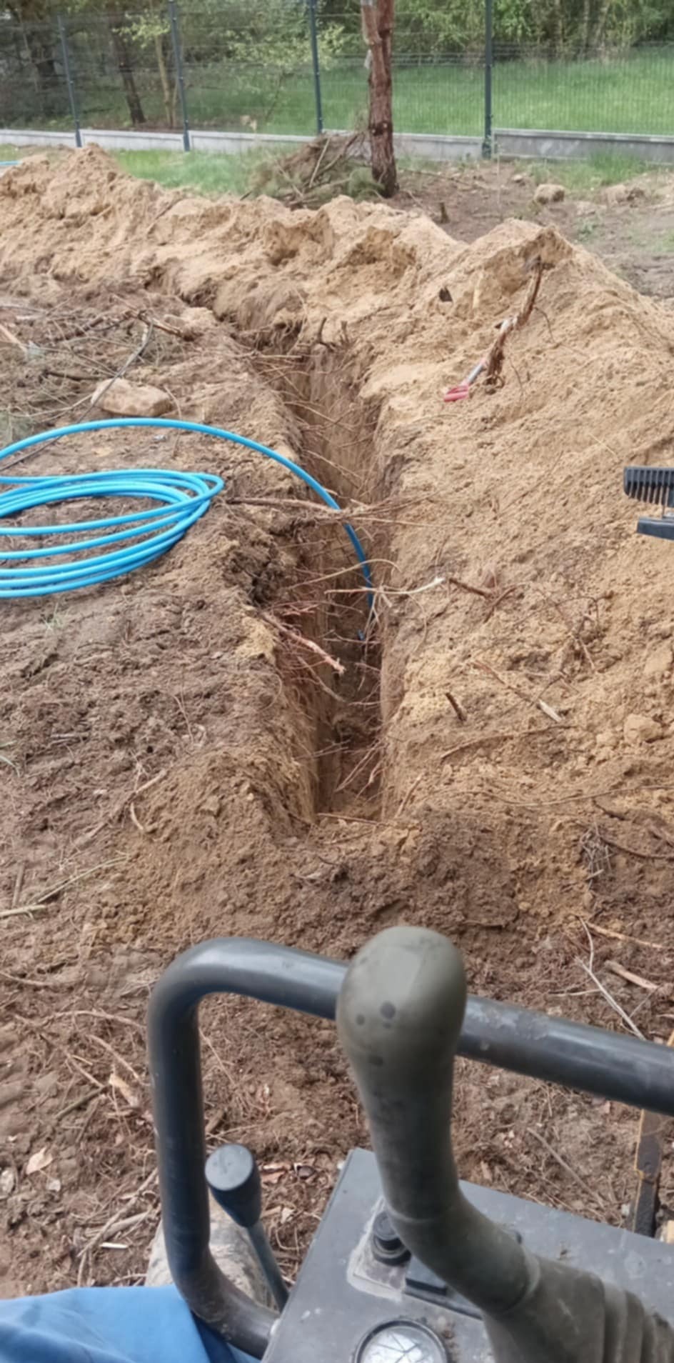 Wąski wykop w ziemi z niebieskim przewodem wewnątrz, widok z perspektywy operatora sprzętu budowlanego. W tle trawa i ogrodzenie.
