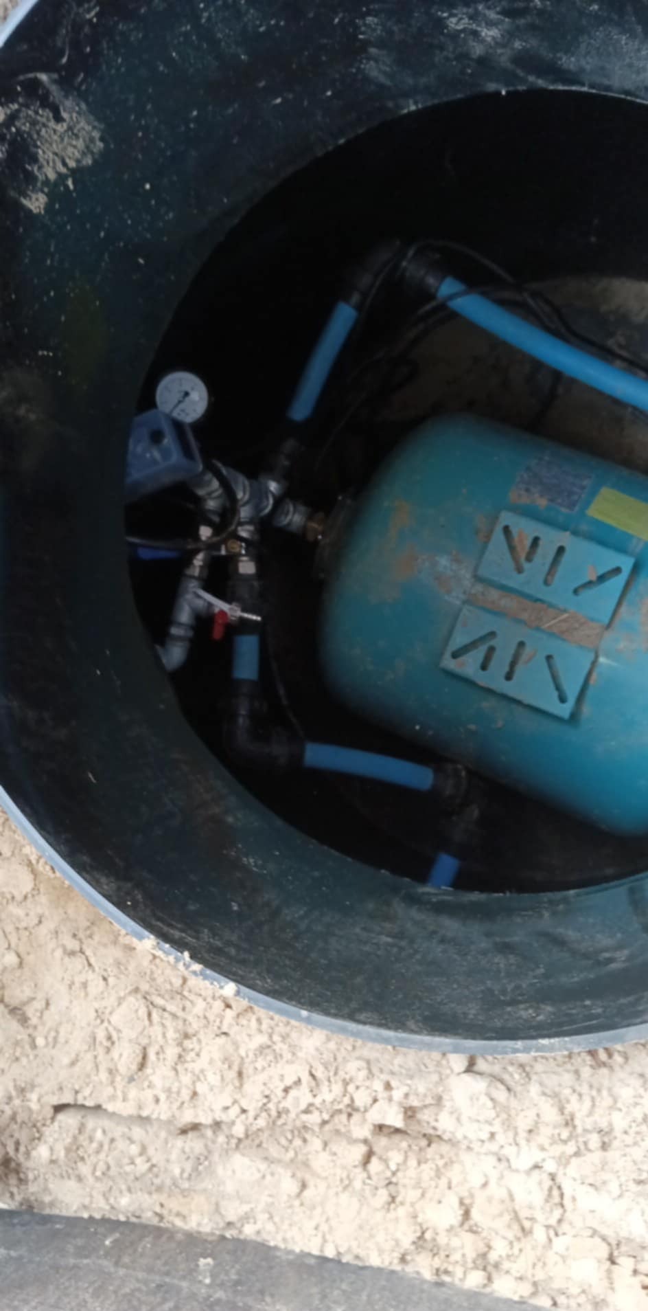Wnętrze studni kręgowej z hydroforem, rurami i manometrem. Widok z góry na zamontowaną instalację w betonowym kręgu studni. Elementy instalacji w kolorze niebieskim i czarnym.
