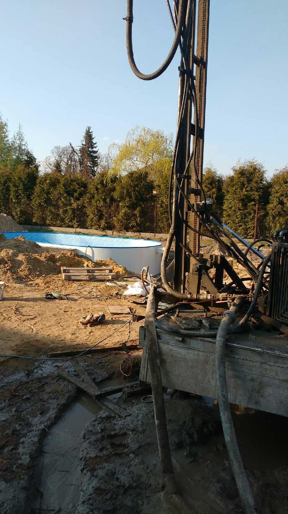 Wiertnica w trakcie pracy, obok częściowo zagłębiony basen stelażowy. Widoczne błoto i teren budowy. Ujęcie z bliska, skupione na maszynie.