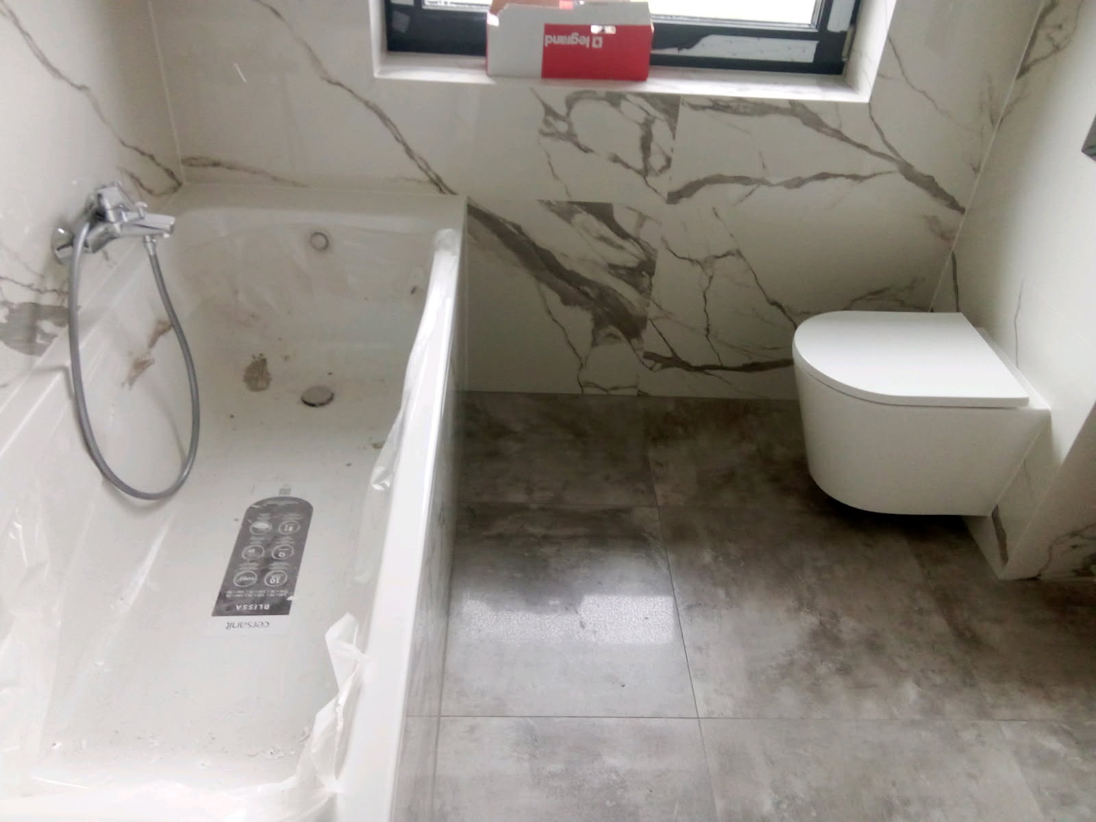 Nowoczesna łazienka z marmurowymi płytkami, białą wanną z baterią i podwieszaną toaletą. Widoczna kartonowa skrzynka na parapecie.