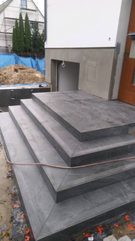 Zewnętrzne schody wejściowe wyłożone ciemnoszarą płytką imitującą kamień, z subtelnym wzorem. Widoczne detale wykończenia krawędzi i fuga. Częściowo widoczny budynek w tle.