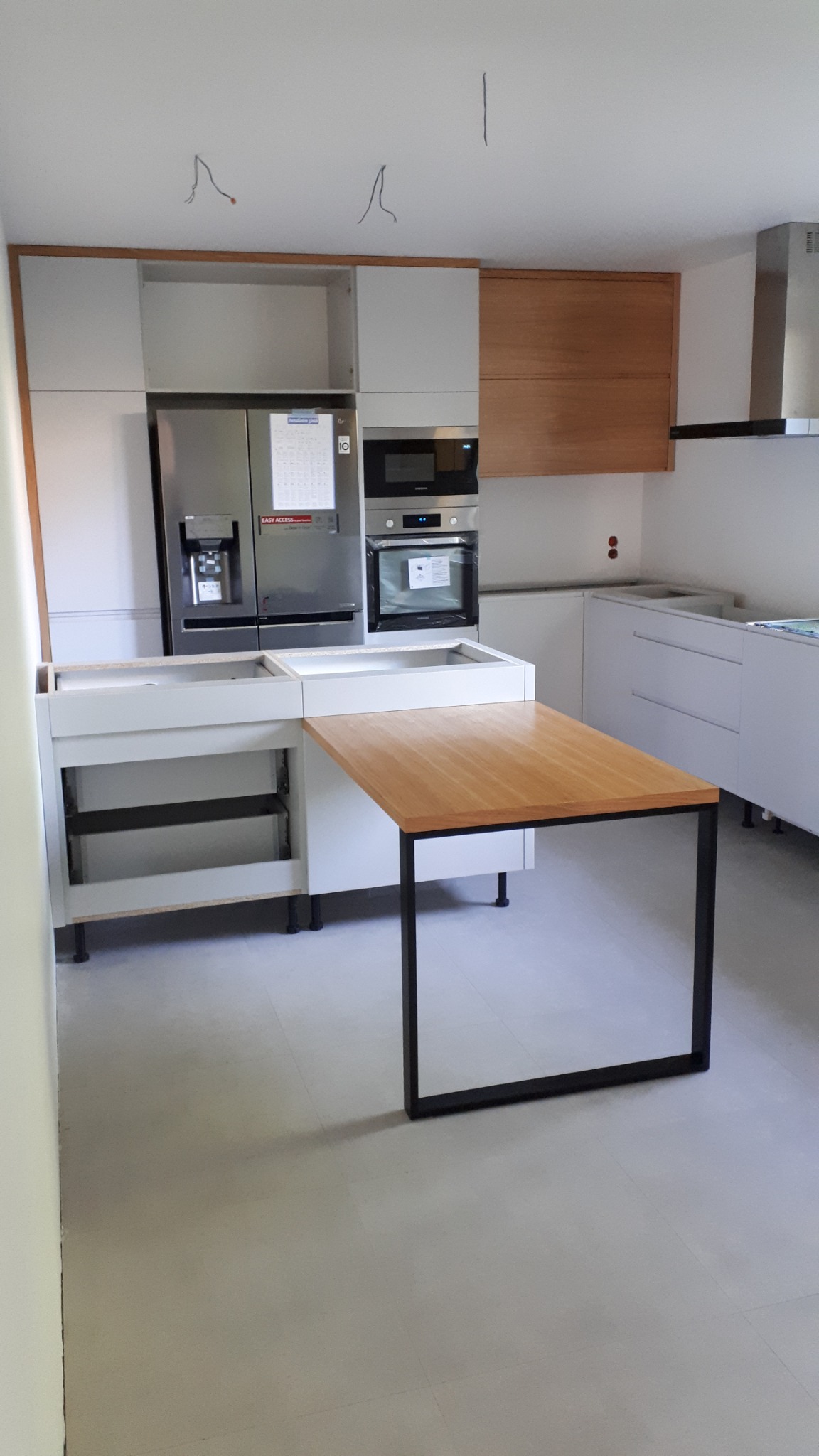 Montaż jasnej kuchni z drewnianymi akcentami: wysokie szafki, sprzęt AGD (lodówka, piekarnik), wyspa z drewnianym blatem na czarnych nogach. Etap prac montażowych.