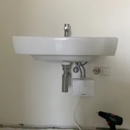 Biała umywalka z baterią i podgrzewaczem wody zamontowana na ścianie. Obok wiertarka i narzędzia hydrauliczne. Widoczne ślady po remoncie.