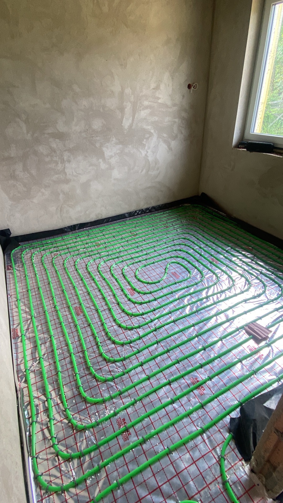 Zielone rury ogrzewania podłogowego ułożone spiralnie na folii z siatką w surowym pomieszczeniu z oknem. Widoczne elementy instalacji i przygotowania do wylewki.