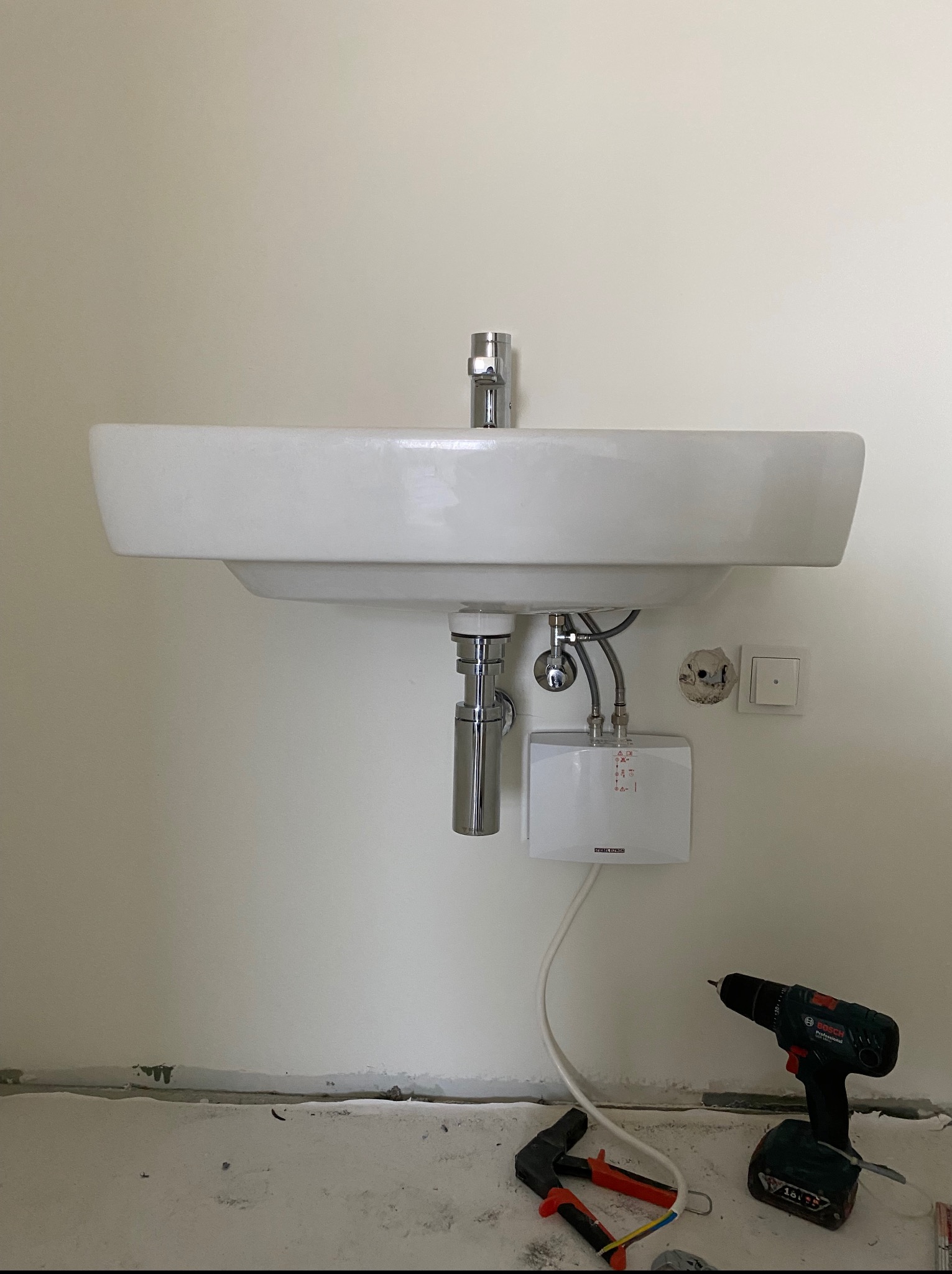 Biała umywalka z baterią i podgrzewaczem wody zamontowana na ścianie. Obok wiertarka i narzędzia hydrauliczne. Widoczne ślady po remoncie.