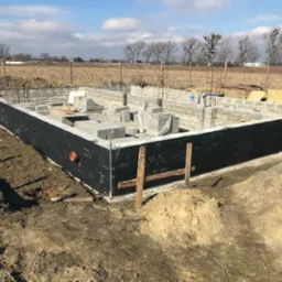 Wznoszenie fundamentów domu z bloczków betonowych, zabezpieczonych czarną izolacją, na tle zaoranego pola i błękitnego nieba.