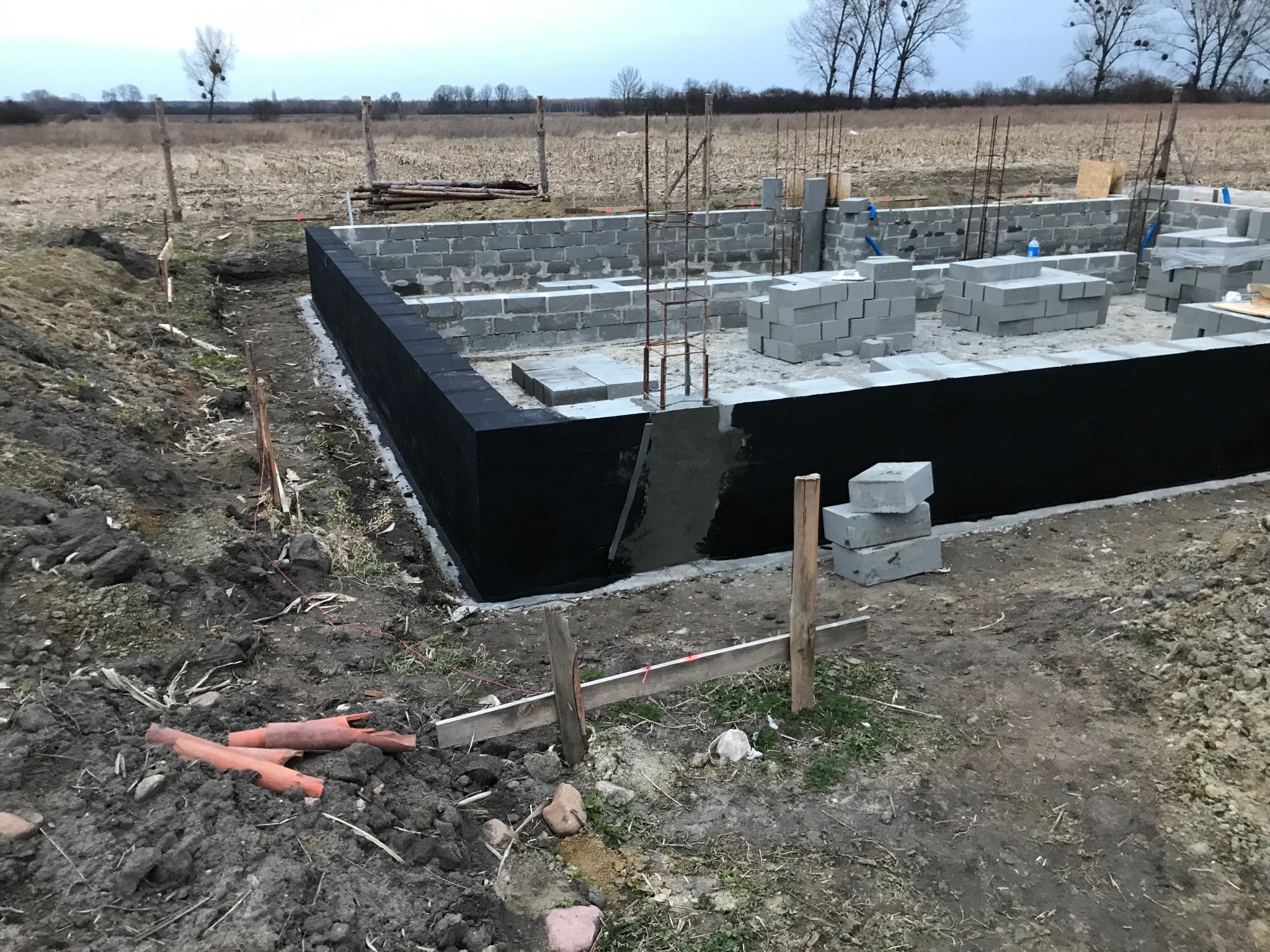 Fundamenty domu z bloczków betonowych, zabezpieczone czarną izolacją, z widocznym zbrojeniem i stertą bloczków na placu budowy. Krajobraz polny w tle.