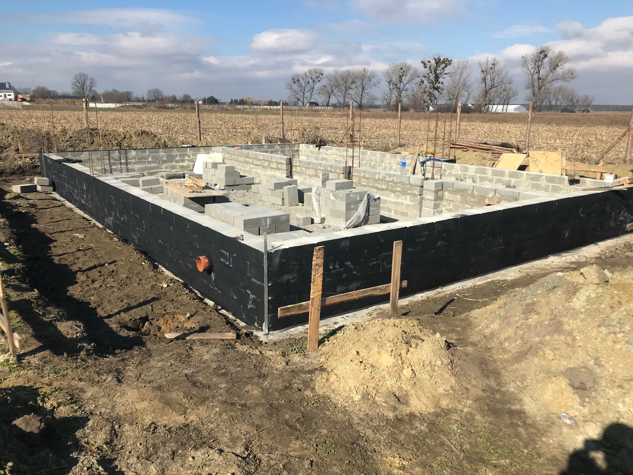 Wznoszenie fundamentów domu z bloczków betonowych, zabezpieczonych czarną izolacją, na tle zaoranego pola i błękitnego nieba.