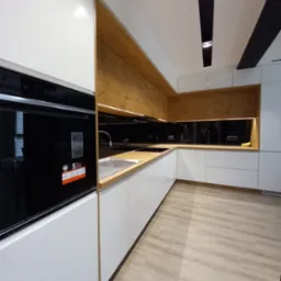 Nowoczesna kuchnia z białymi frontami i drewnianymi akcentami. Widoczny piekarnik z wyświetlaczem, drewniany blat i czarny panel nad nim. Oświetlenie podszafkowe.