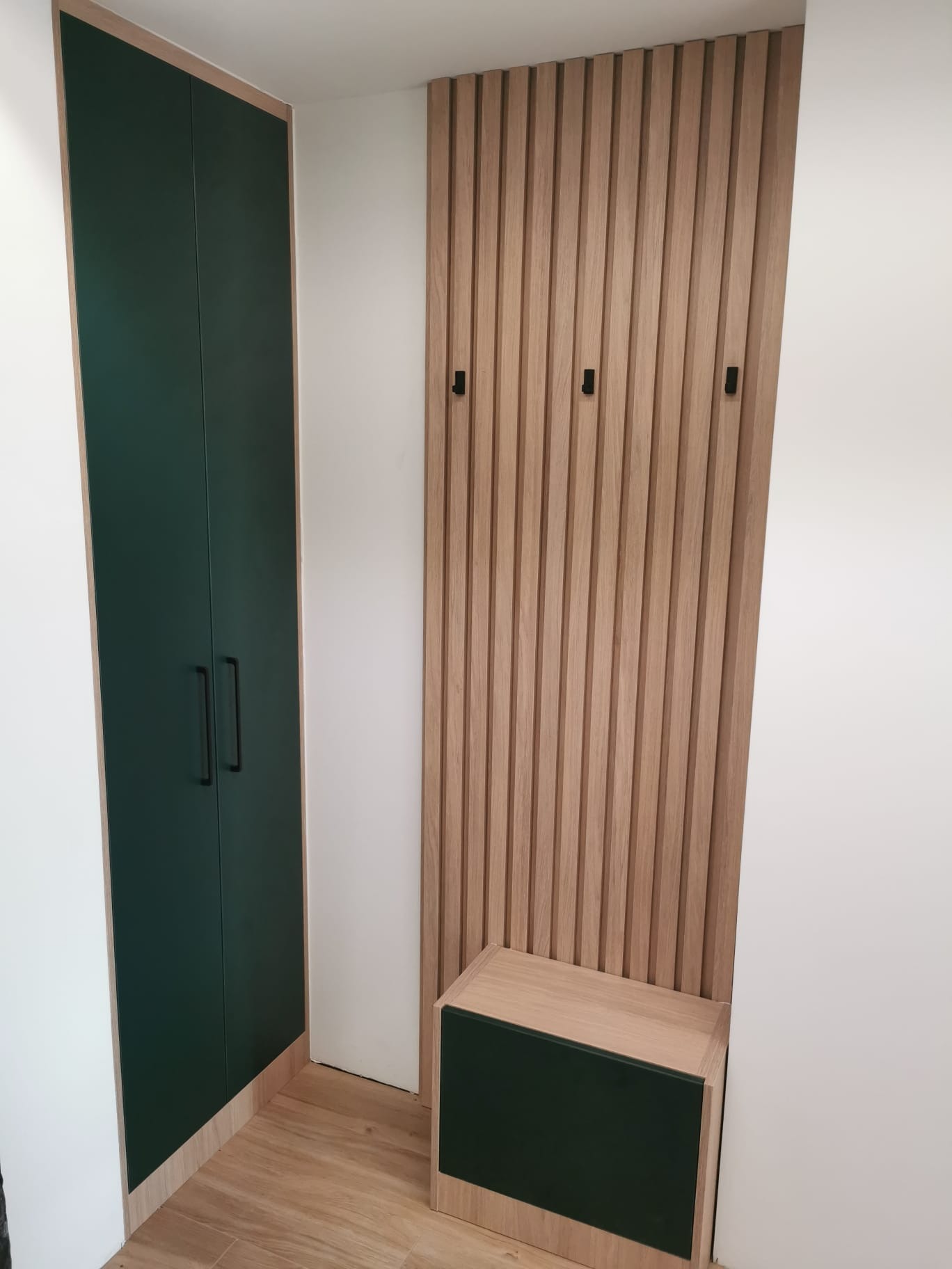 Nowoczesna zabudowa przedpokoju w Tychach: ciemnozielona szafa wnękowa z czarnymi uchwytami i panel z drewnianych listew z wieszakami oraz siedzisko.