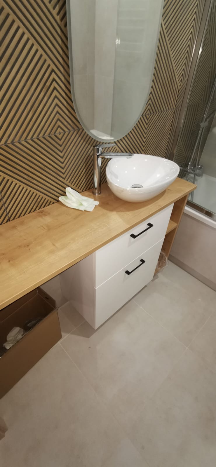 Nowoczesna łazienka z drewnianym blatem, białą umywalką nablatową i szafką z czarnymi uchwytami. Na ścianie dekoracyjne panele w geometryczny wzór.