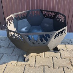 TopMetalWork - Czarny, metalowy kosz na ognisko z wyciętymi motywami płomieni stoi na kostce brukowej. W tle blaszane ogrodzenie.