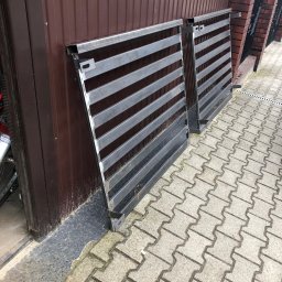 TopMetalWork - Dwie surowe, metalowe bramy wjazdowe z poziomymi szczeblami, oparte o brązową ścianę i mur z czerwonej cegły na kostce brukowej. Widoczne elementy montażowe.