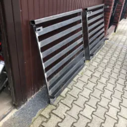 Dwie surowe, metalowe bramy wjazdowe z poziomymi szczeblami, oparte o brązową ścianę i mur z czerwonej cegły na kostce brukowej. Widoczne elementy montażowe.