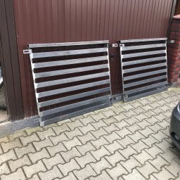 TopMetalWork - Dwie metalowe bramy wjazdowe z poziomymi prętami, przygotowane do montażu przed brązową bramą garażową i obok zaparkowanego, brudnego samochodu.