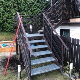TopMetalWork - Zewnętrzne, metalowe schody z ażurowymi stopniami prowadzące do drewnianej konstrukcji, z elementami ogrodu w tle i czerwoną miarką opartą o poręcz.
