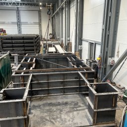 TopMetalWork - Wnętrze hali produkcyjnej z konstrukcją metalową w trakcie budowy. Widoczny pracownik w kombinezonie i butla z gazem. Elementy konstrukcji podnoszone dźwigiem.