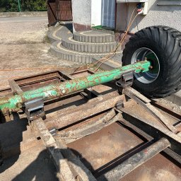 TopMetalWork - Metalowa konstrukcja z dużymi kołami, prawdopodobnie podwozie przyczepy rolniczej, z widocznymi spawami i śladami rdzy, ustawiona na zewnątrz budynku.