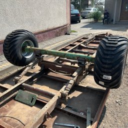 TopMetalWork - Rama metalowa z dużymi kołami, prawdopodobnie w trakcie budowy przyczepy. Widoczne narzędzia ślusarskie i elementy konstrukcyjne. Tło stanowi budynek mieszkalny.