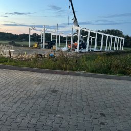 TopMetalWork - Wznoszenie stalowej konstrukcji hali z użyciem dźwigu na tle zachodzącego słońca. Widoczne elementy szkieletu i maszyny budowlane.