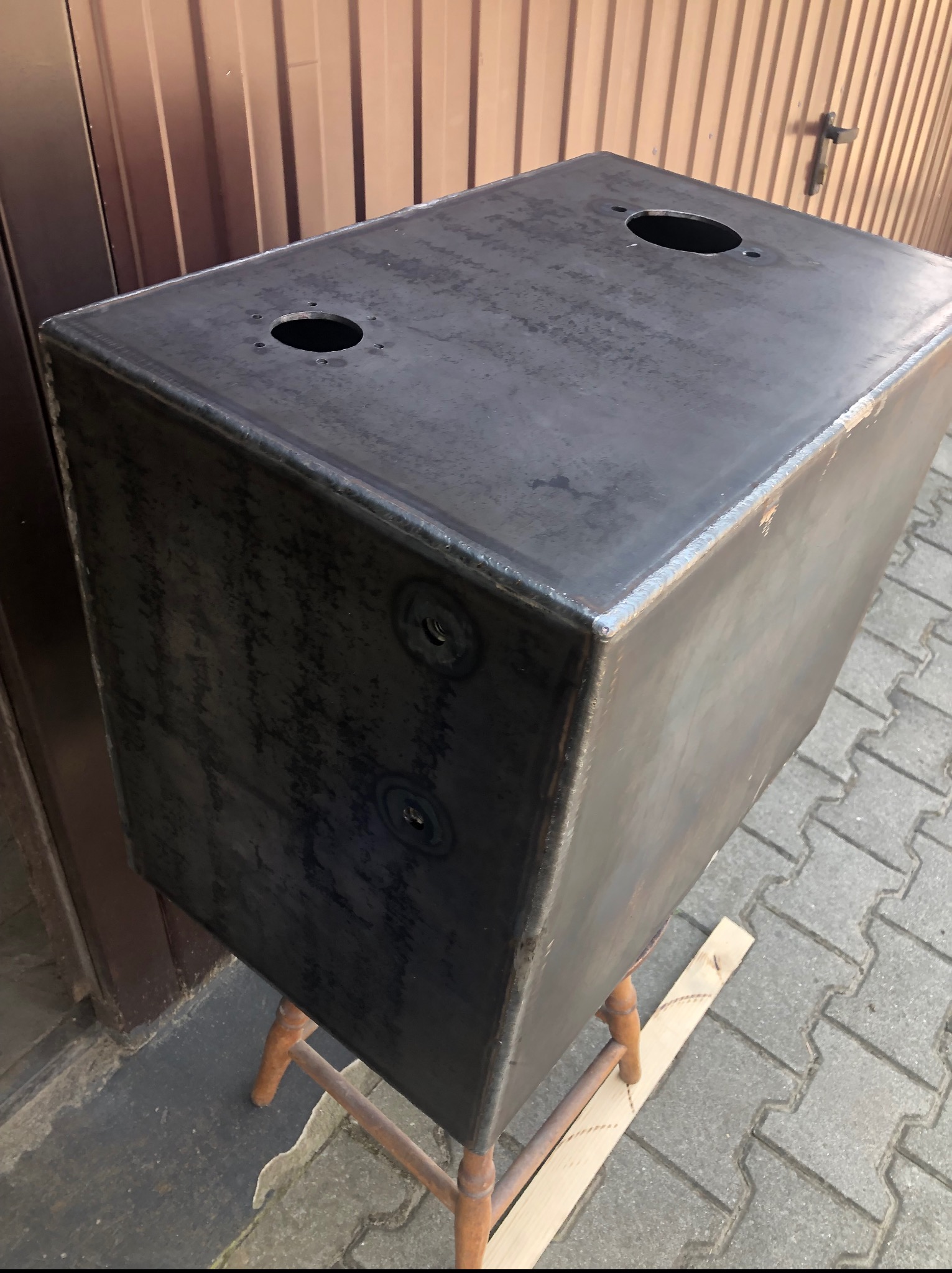 Czarny, metalowy zbiornik z otworami na górze, spawany na krawędziach, stoi na drewnianym stołku na kostce brukowej, widok z góry pod kątem.