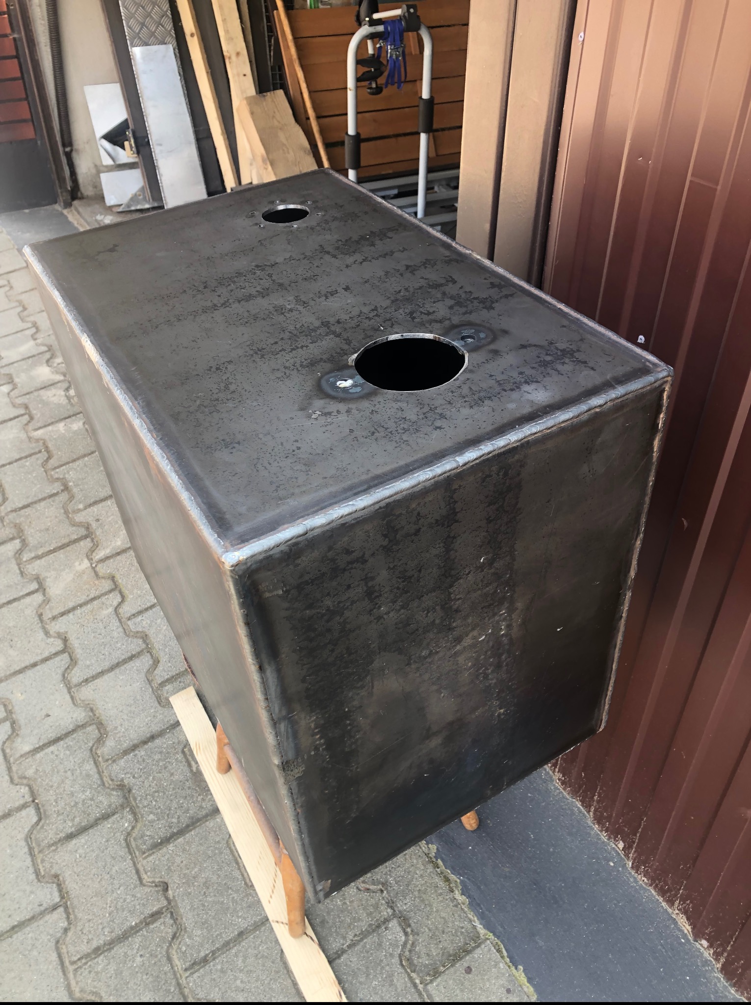 Spawana, metalowa konstrukcja skrzyniowa z otworami na górze, postawiona na drewnianych elementach. Widoczne spawy i surowa powierzchnia metalu.