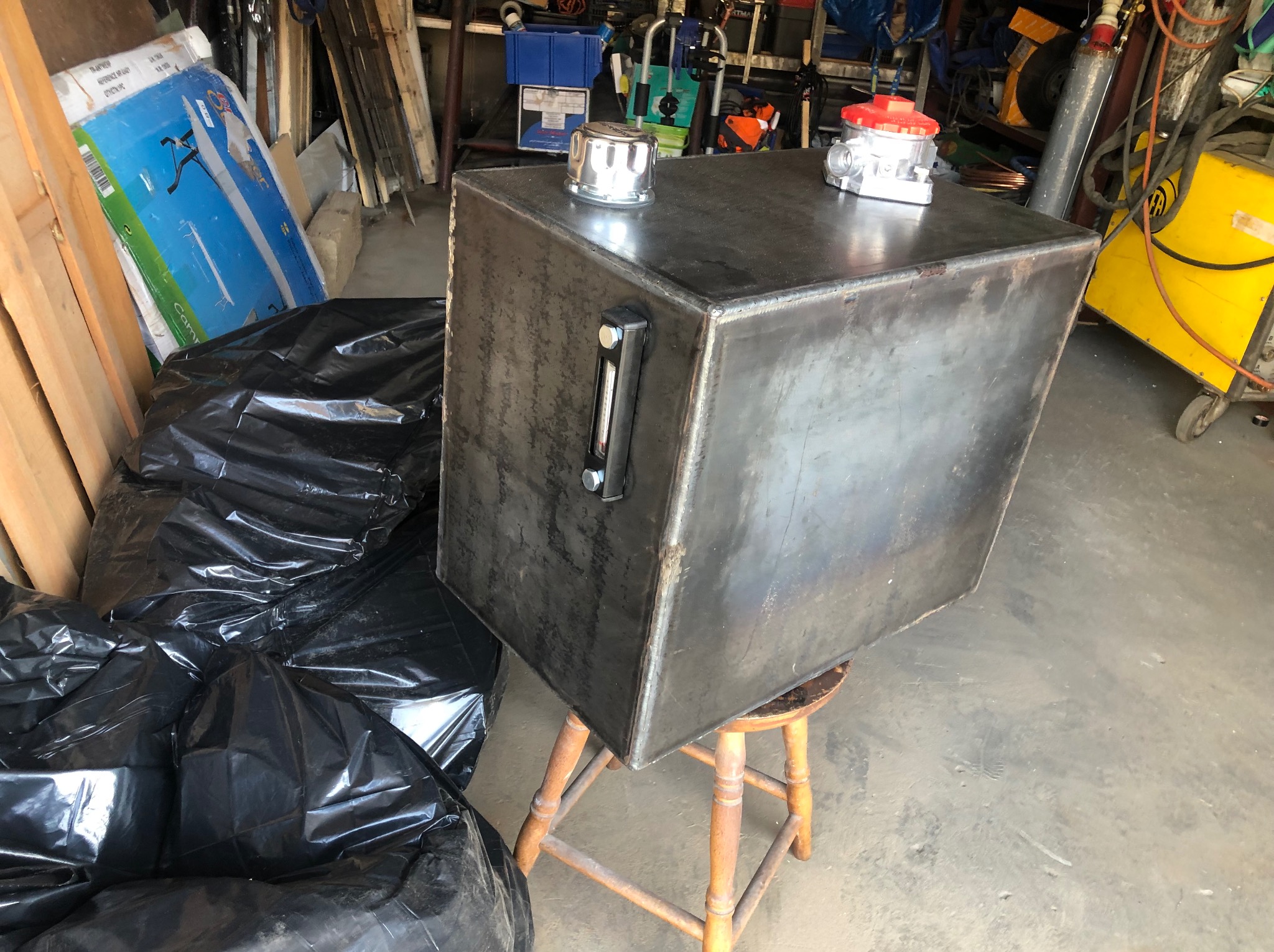 Metalowy zbiornik z chromowanym wlewem i wskaźnikiem poziomu cieczy, umieszczony na drewnianym stołku. W tle widoczny sprzęt warsztatowy, butla gazowa i worki na śmieci.