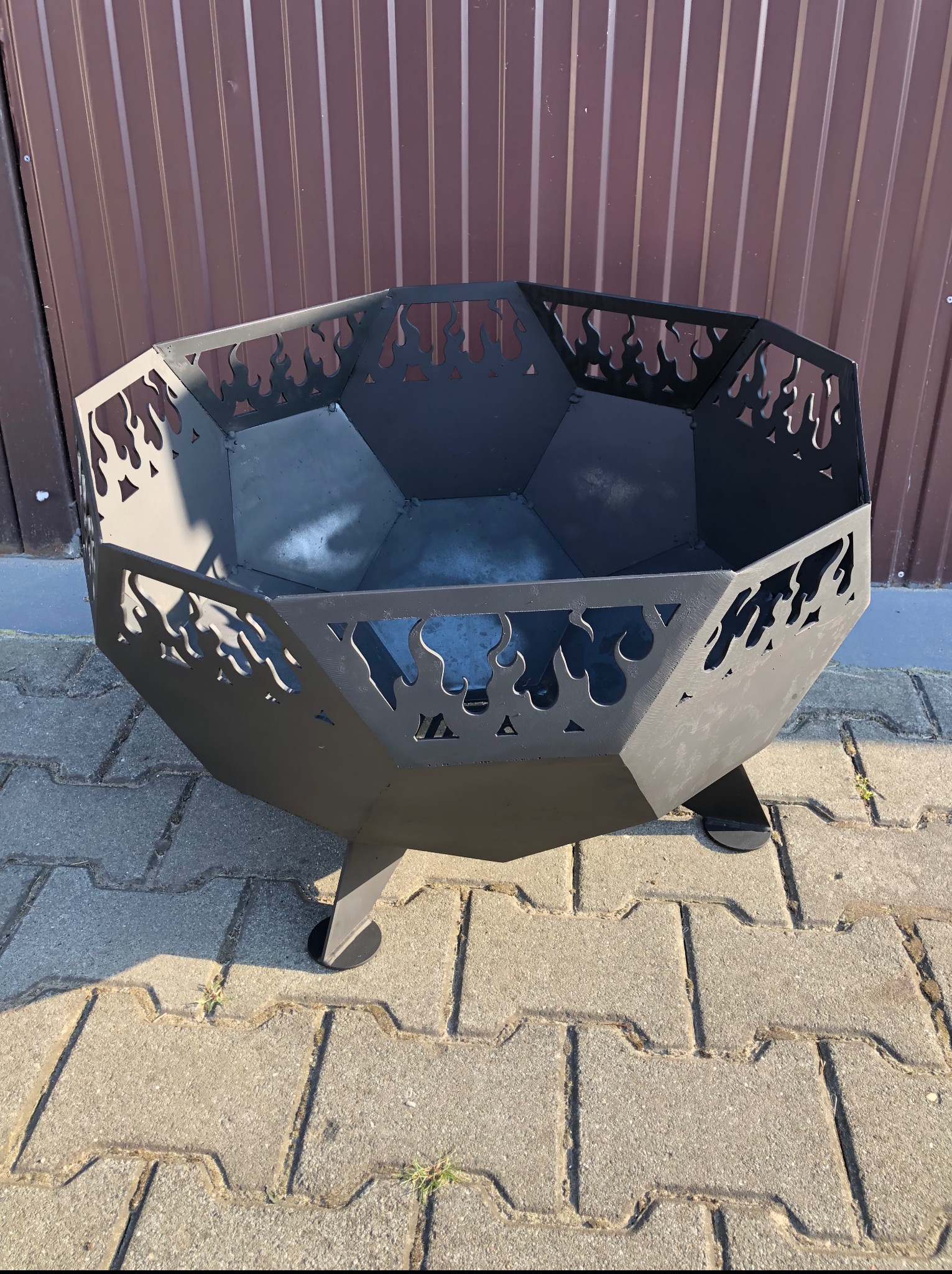 Czarny, metalowy kosz na ognisko z wyciętymi motywami płomieni stoi na kostce brukowej. W tle blaszane ogrodzenie.