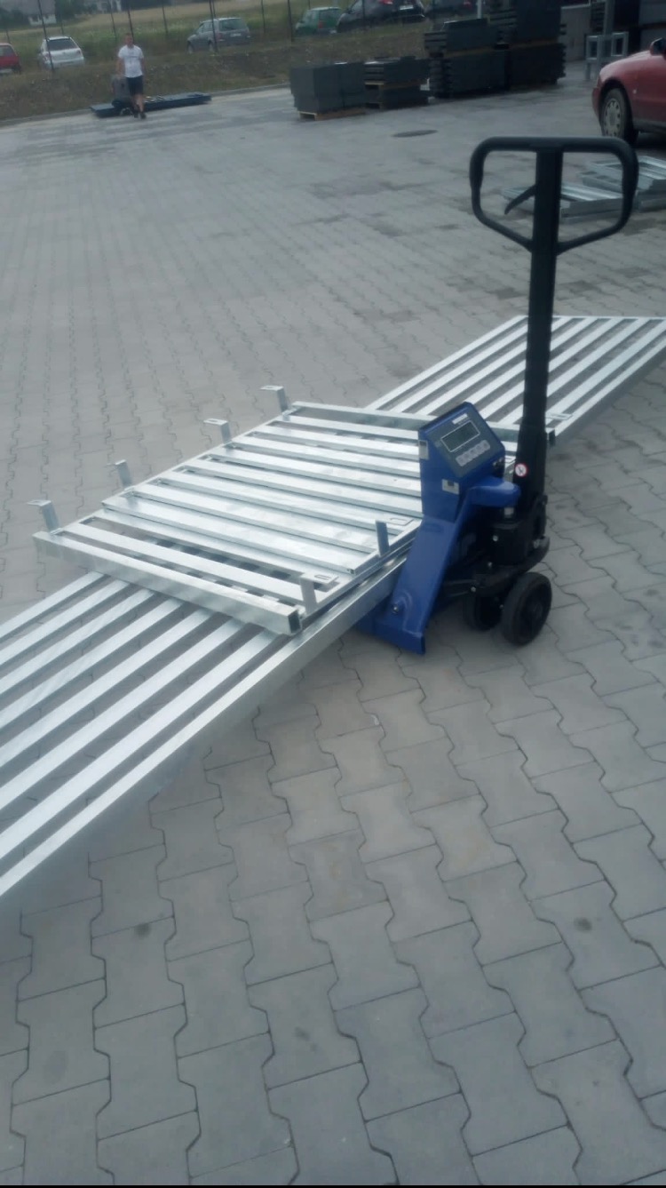 Wózek paletowy z ładunkiem ocynkowanych elementów konstrukcyjnych na zewnątrz, prawdopodobnie fragmentów ogrodzenia lub balustrady, na brukowanym placu.