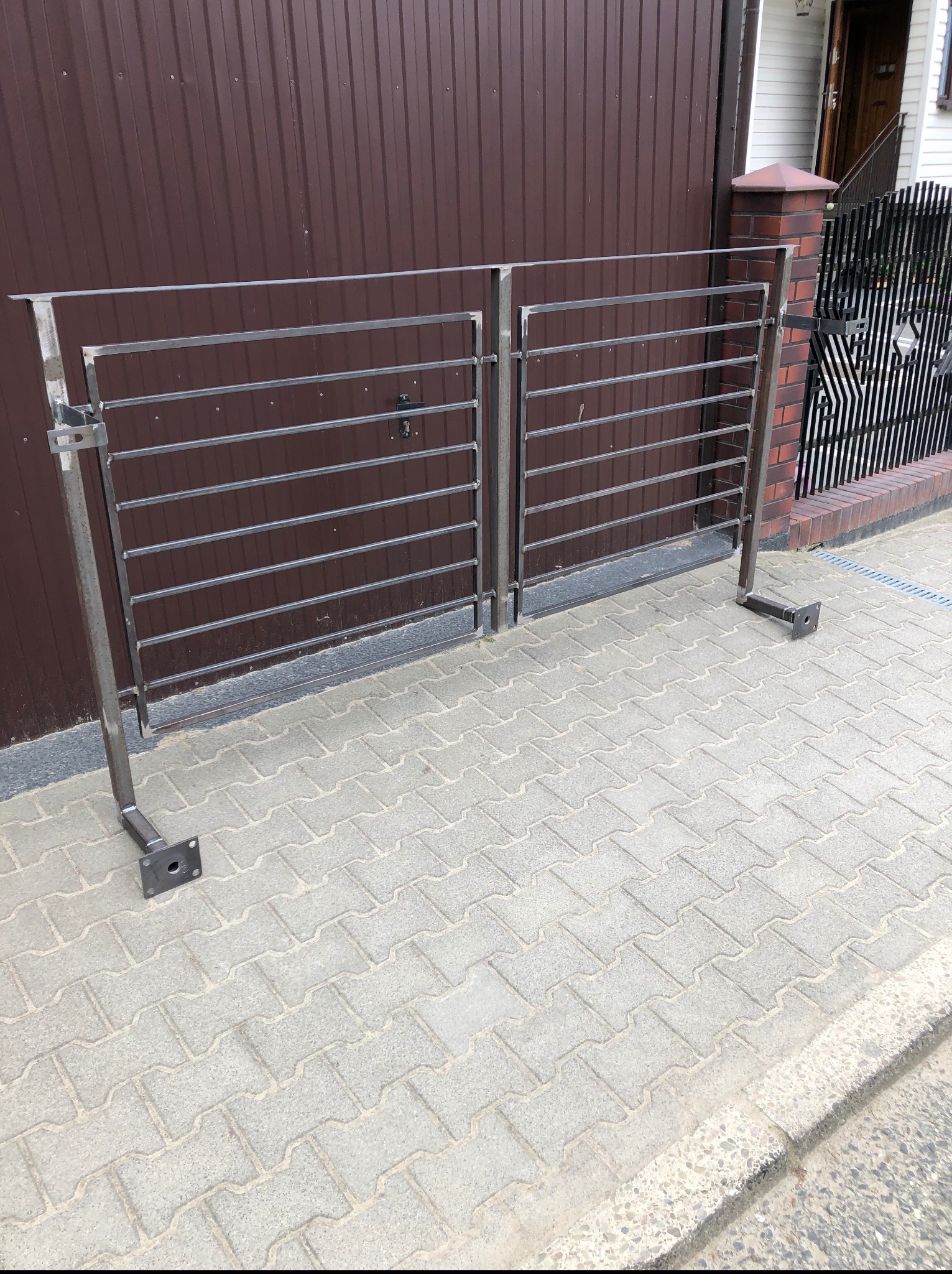 Surowa, metalowa brama dwuskrzydłowa z poziomymi prętami, bez wykończenia, z widocznymi mocowaniami do słupków, na tle brązowej blachy i kostki brukowej.