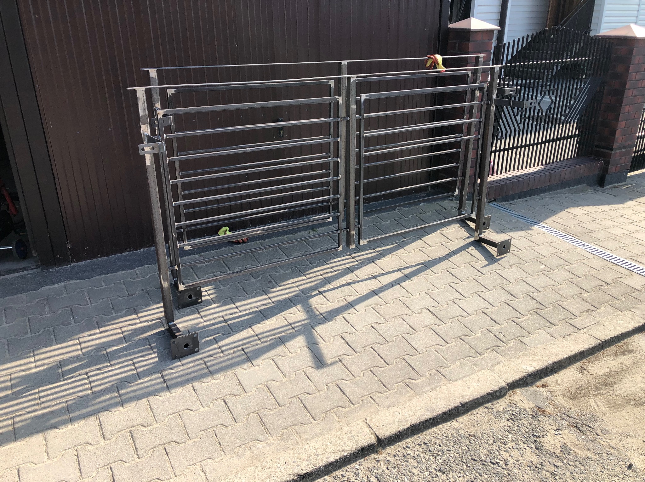 Surowa, metalowa konstrukcja dwuskrzydłowej bramy wjazdowej z poziomymi szczeblami, przygotowana do montażu na posesji z brązową bramą.