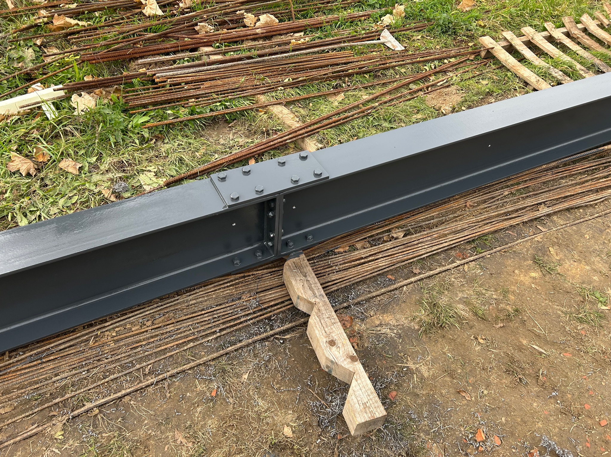 Połączony, pomalowany na szaro profil stalowy na ziemi z prętami zbrojeniowymi i drewnianym podparciem. Widoczne ślady obróbki metalu.