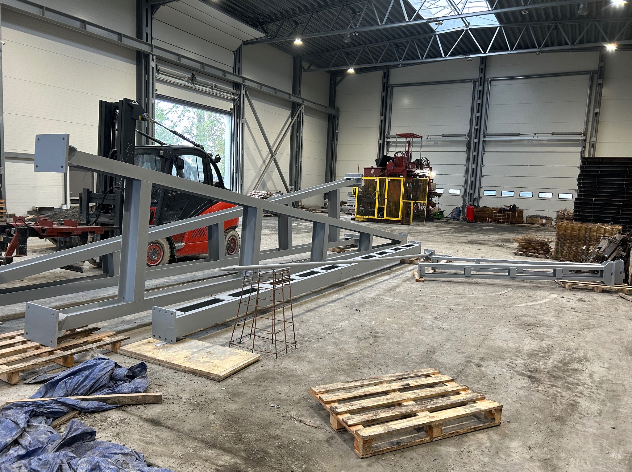 Szare, metalowe elementy konstrukcyjne w hali produkcyjnej, z wózkiem widłowym w tle. Widoczne palety i maszyny, surowe wykończenie wnętrza.