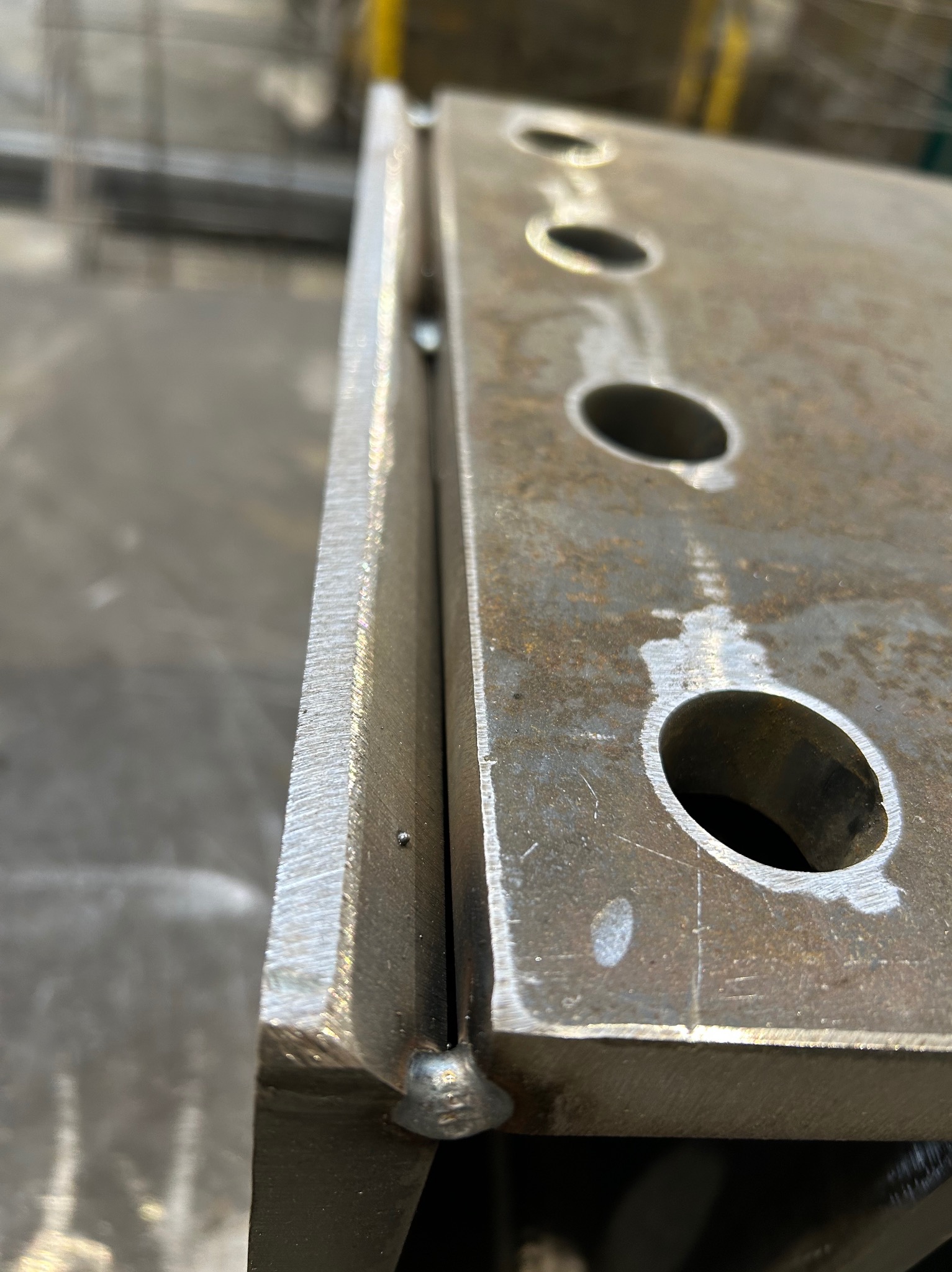 Spoina łącząca dwie metalowe płyty z otworami, widoczna z bliska. Szlifowana krawędź i ślady po obróbce. Detal konstrukcji stalowej w hali.