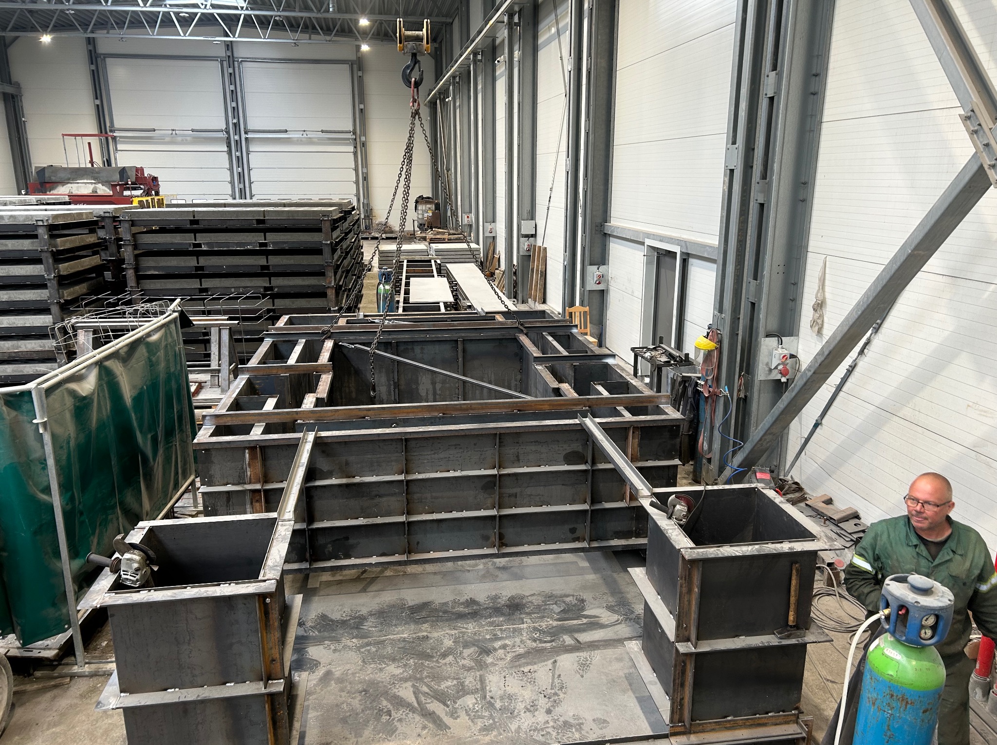 Wnętrze hali produkcyjnej z konstrukcją metalową w trakcie budowy. Widoczny pracownik w kombinezonie i butla z gazem. Elementy konstrukcji podnoszone dźwigiem.