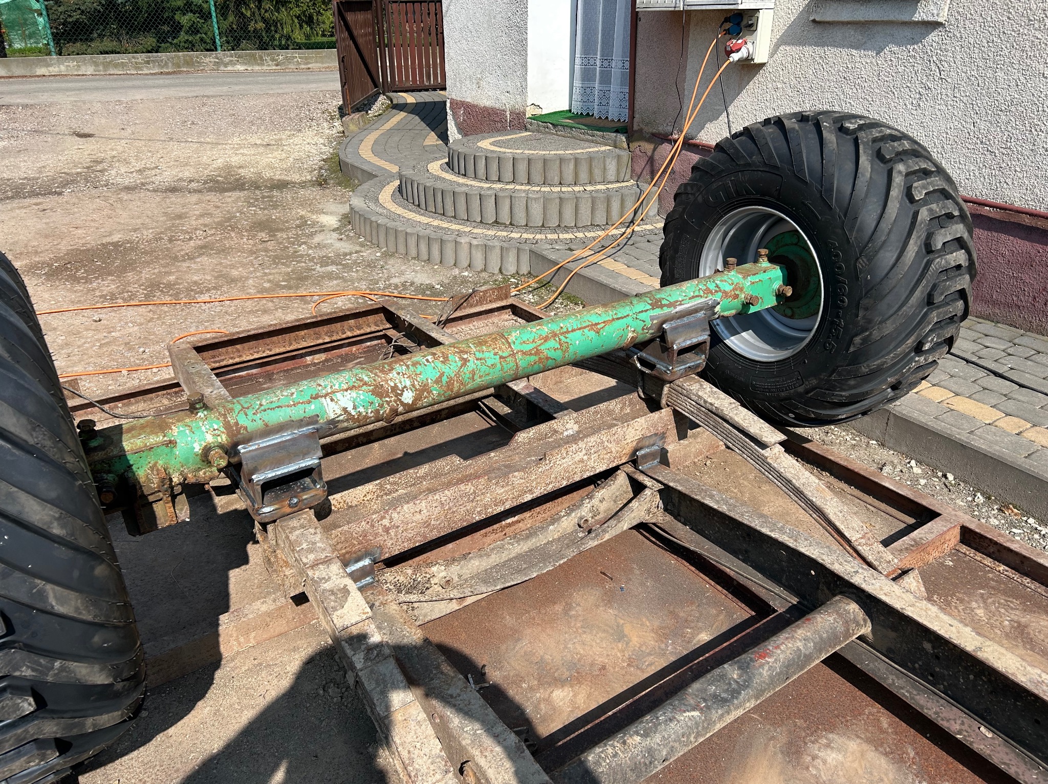 Metalowa konstrukcja z dużymi kołami, prawdopodobnie podwozie przyczepy rolniczej, z widocznymi spawami i śladami rdzy, ustawiona na zewnątrz budynku.