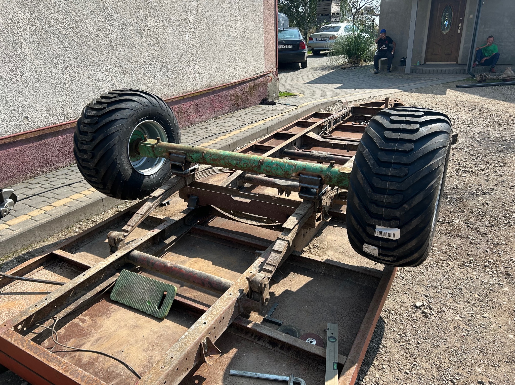 Rama metalowa z dużymi kołami, prawdopodobnie w trakcie budowy przyczepy. Widoczne narzędzia ślusarskie i elementy konstrukcyjne. Tło stanowi budynek mieszkalny.
