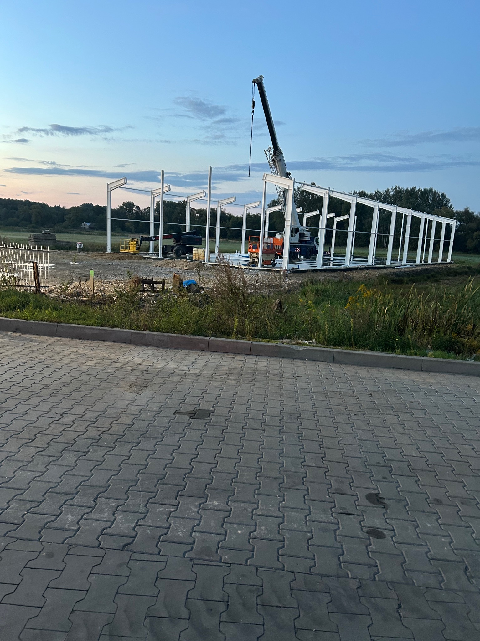 Wznoszenie stalowej konstrukcji hali z użyciem dźwigu na tle zachodzącego słońca. Widoczne elementy szkieletu i maszyny budowlane.