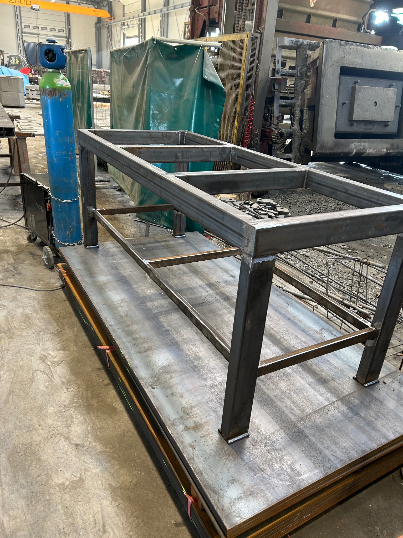 Stalowa konstrukcja stołu warsztatowego w trakcie spawania, widoczny spawany szkielet na stosie blach, butla z gazem i spawarka w tle hali produkcyjnej.