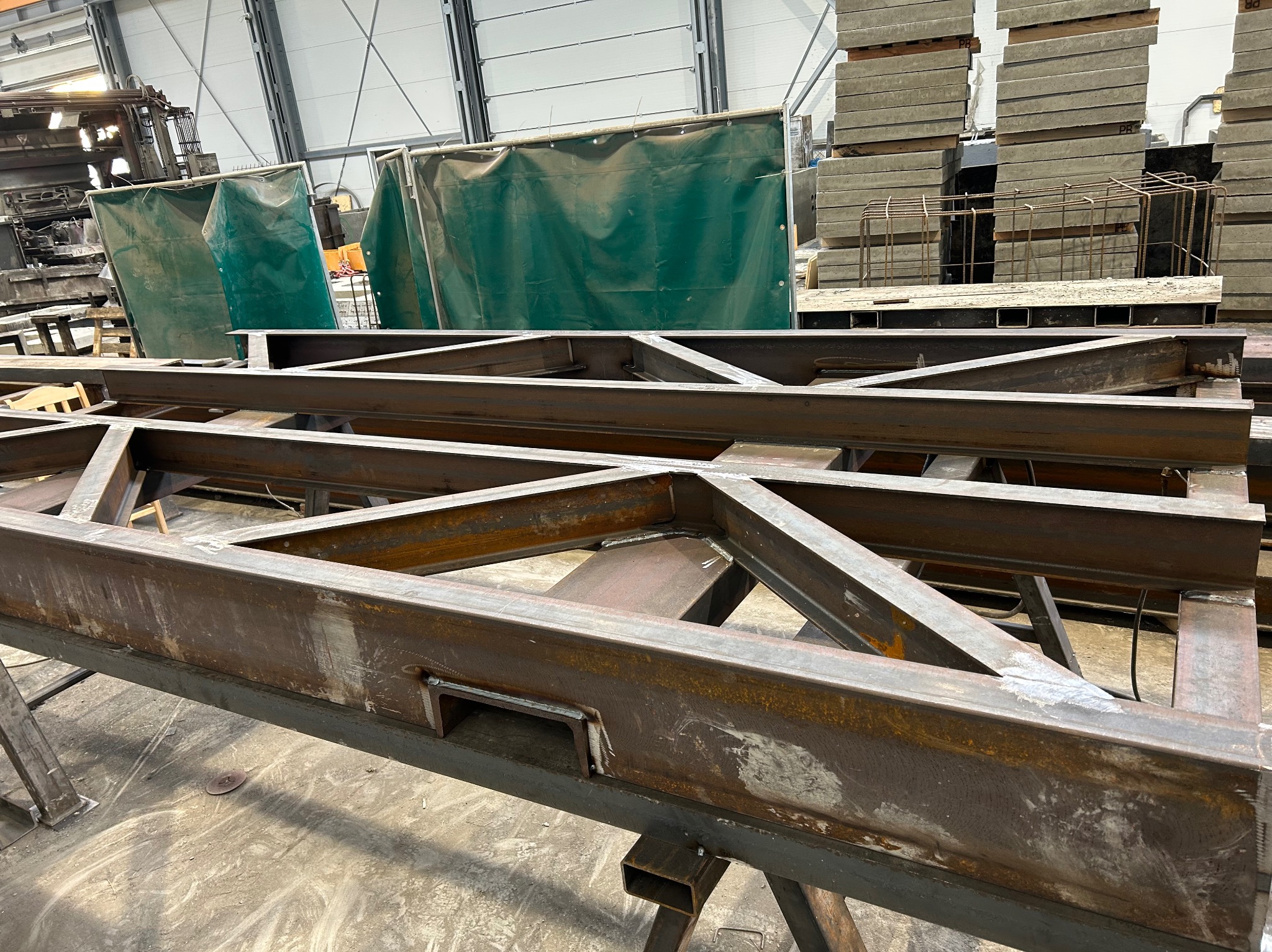 Konstrukcja stalowa z profili, spawana, widoczne ślady obróbki, w tle elementy betonowe i osłony spawalnicze. Surowy, industrialny charakter.