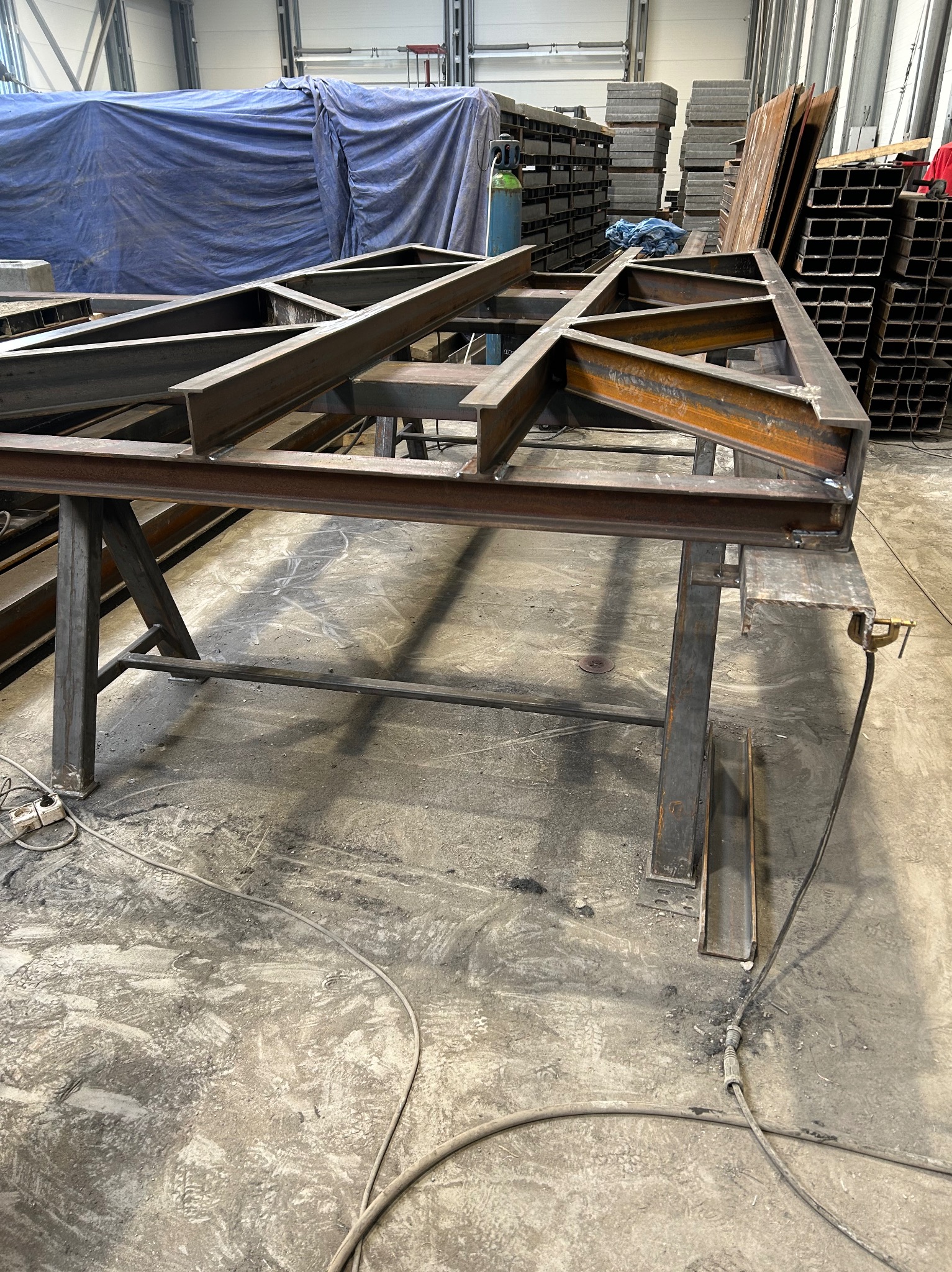 Konstrukcja metalowa z profili spawanych na stole warsztatowym, widoczne ślady spawania, w tle materiały i narzędzia w hali produkcyjnej. Surowy, industrialny klimat.