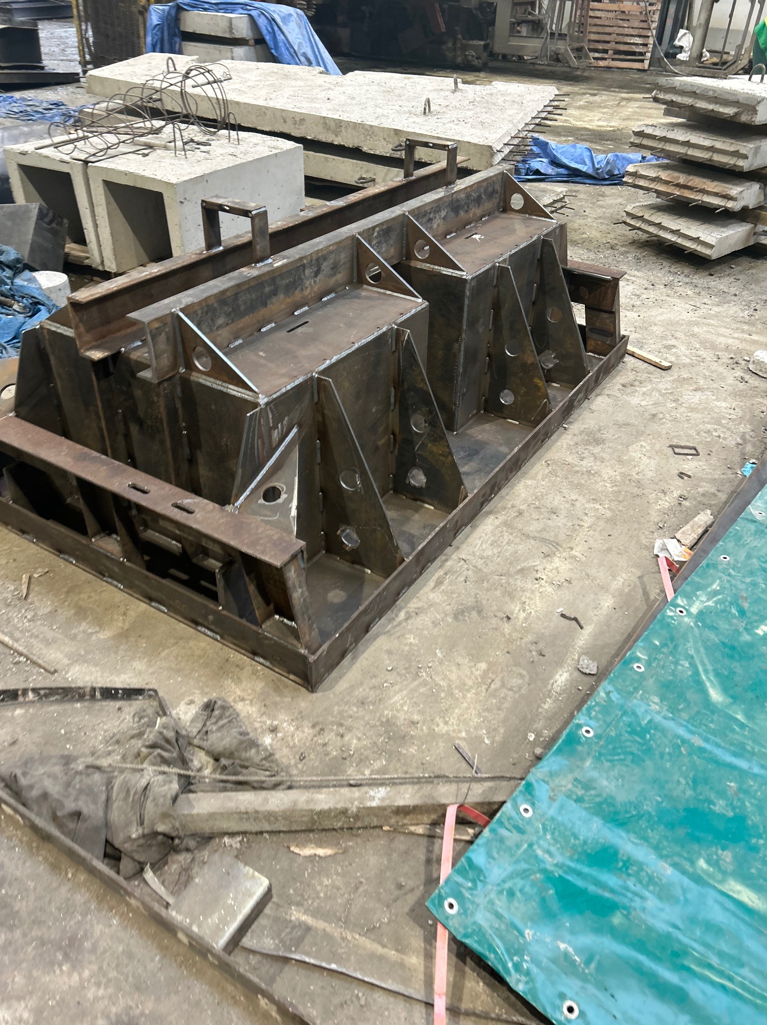 Metalowa forma do odlewania betonu z widocznymi spawami i otworami, leżąca na betonowej posadzce w hali produkcyjnej. W tle elementy betonowe.