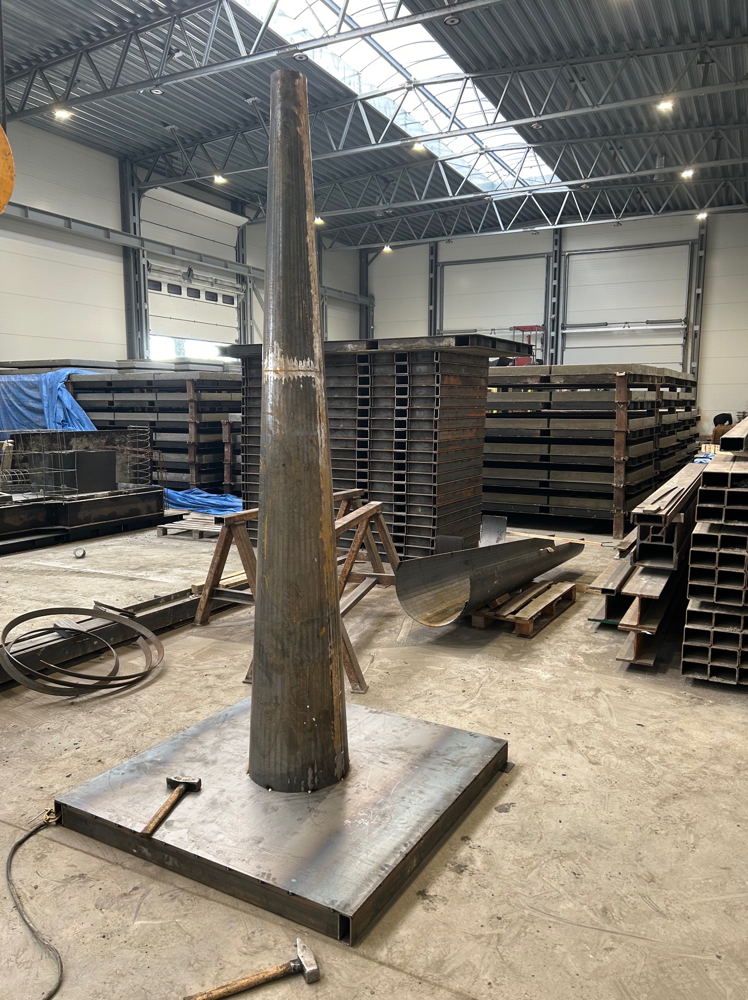 Metalowa konstrukcja w hali produkcyjnej, spawana kolumna na podstawie, w tle stosy profili i blach. Widoczne narzędzia ślusarskie.