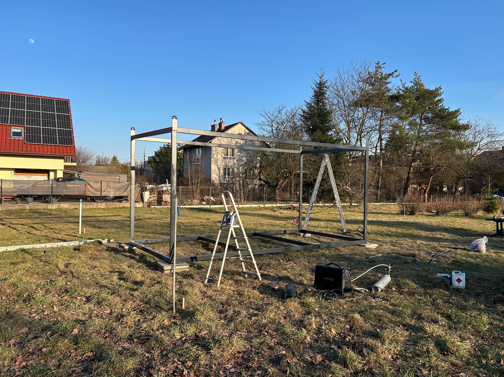 Metalowa konstrukcja szkieletowa na trawniku w słoneczny dzień, z widocznymi narzędziami spawalniczymi i drabinami, sugerująca etap budowy w otoczeniu domów i drzew.