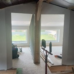 Stanisław szymulewski - Montaż ścianki działowej z płyt gipsowo-kartonowych na poddaszu, widoczne narzędzia i materiały budowlane, okna w tle.